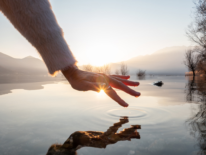 Detail of hand touching and caressing water surface of beautiful lake at sunset, mountain view. Purity freshness clean concept, one person touching lake with hand