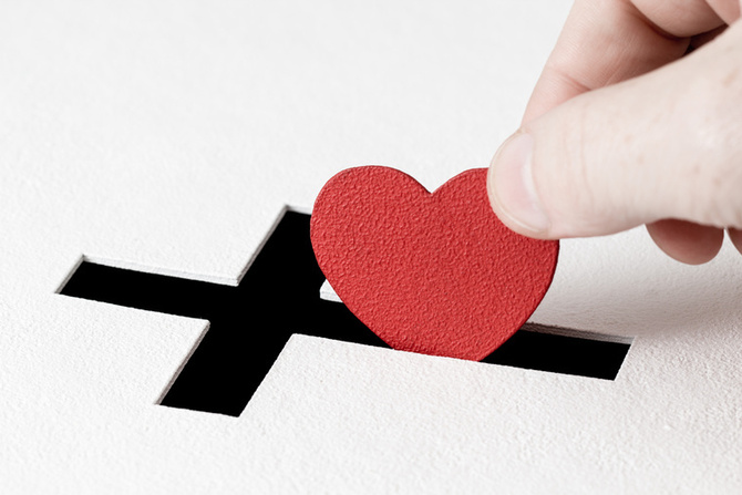 Hand of parishioner is inserting heart symbol into hole for donations in form of Christian cross on white background. Idea of sincere devotion for faith with all heart
