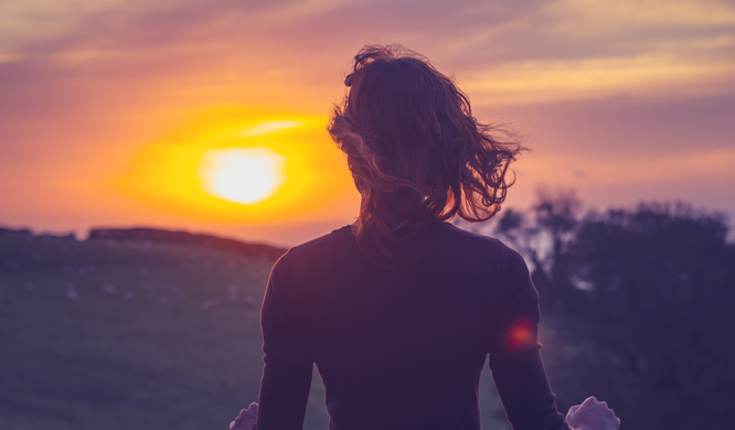 Rear view of young woman admiring the sunset over a field from her balcony